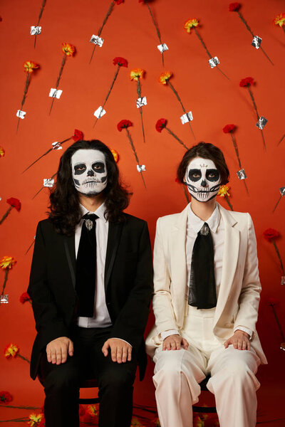 scary dia de los muertos couple sitting on chairs and looking at camera on red floral backdrop