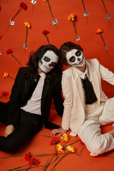 couple in scary catrina makeup and suits sitting on floor in red studio with carnation flowers