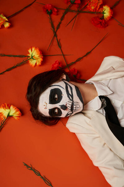 woman in dia de los muertos makeup and suit lying down on red backdrop with flowers, top view