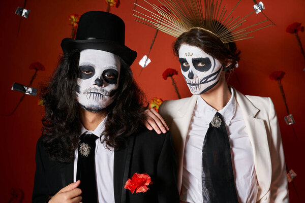 trendy couple in dia de los muertos skull makeup looking at camera on red backdrop with carnations