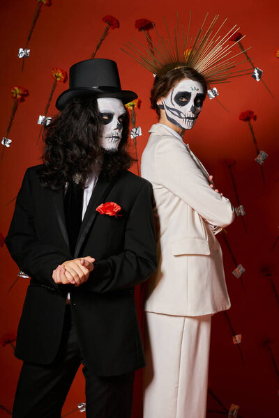 couple in dia de los muertos makeup and festive attire standing back to back on red floral backdrop