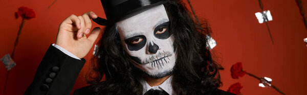 extravagant man in Day of Dead makeup touching top hat on red backdrop with carnations, banner