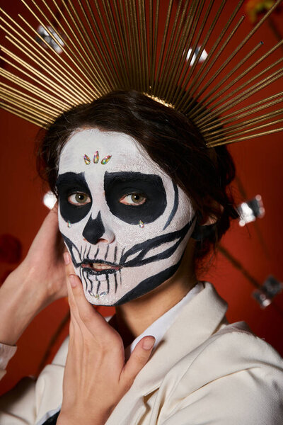 portrait of woman in dia de los muertos makeup and crown looking at camera on red backdrop