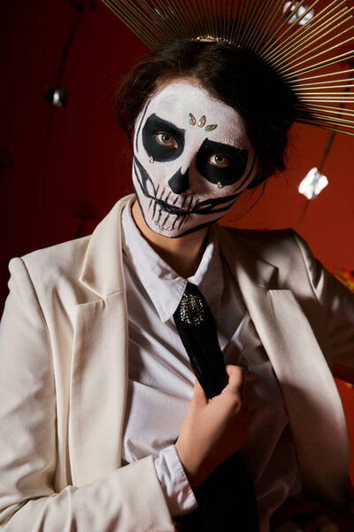 portrait of woman in skeleton makeup and white suit looking at camera on red backdrop, Day of Dead