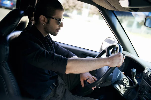 stock image charming man with ponytail in black trendy outfit behind steering wheel of his car, sexy driver