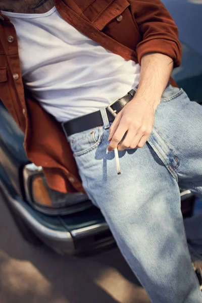 青いジーンズと茶色のシャツの若者の視界が車の隣にタバコを握っている — ストック写真