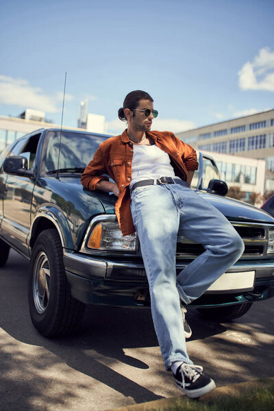 good looking sexy man in vibrant stylish clothes leaning on his car looking away, fashion and style