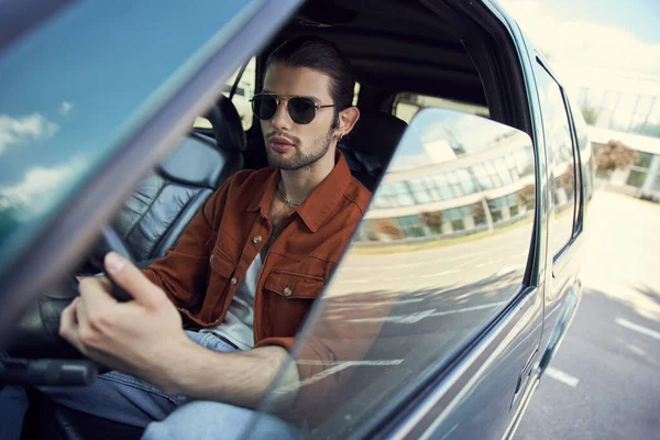handsome young male model in stylish brown shirt with sunglasses at steering wheel, sexy driver