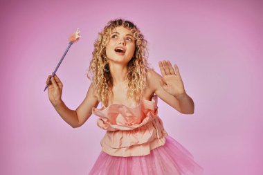 blonde curly haired woman with magic wand posing in pink tooth fairy costume on pink backdrop clipart