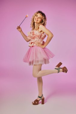 cheerful blonde woman with curly hair in tooth fairy costume standing on one leg on pink backdrop clipart
