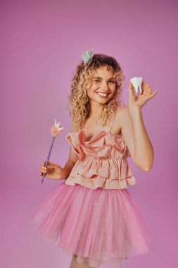 cheerful lovely woman in tooth fairy costume holding magic wand and baby tooth and smiling at camera clipart