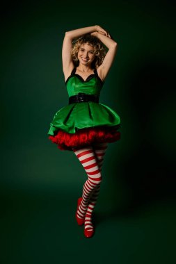 Yeni yıl elfi gibi giyinmiş neşeli bir kadın. Kolunu kaldırıp kameraya gülümsüyor.