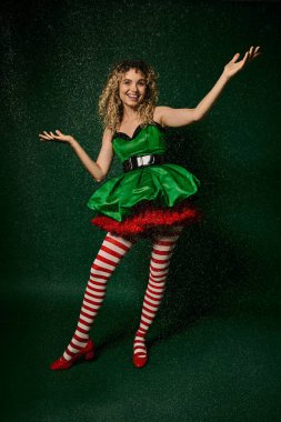 attractive curly woman in festive dress catching snowflakes looking at camera, new year elf concept clipart