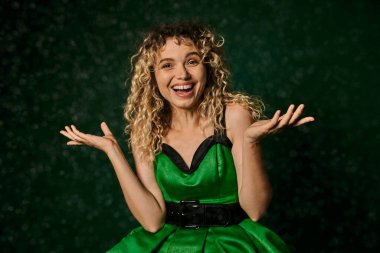 joyous curly woman in new year elf costume standing under falling snow on dark green background clipart