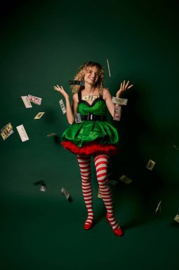 cheerful curly woman dressed as new year elf standing under money rain cheerfully looking at camera clipart