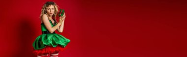 astonished new year elf in green dress with pouted lips holding gift and looking at camera, banner