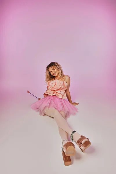 lovely joyous tooth fairy in pink costume sitting on floor with legs crossed on pink backdrop