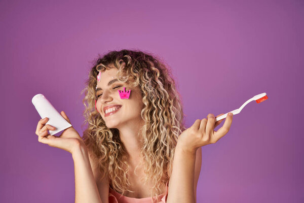 portrait of happy blonde woman with curly hair and face stickers holding toothbrush and paste