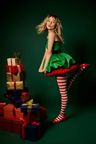 stock image cheerful curly new year elf standing on one leg in green festive dress next to pile of presents