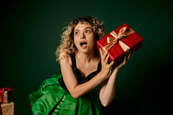 amazed blonde new year elf posing in green dress with present in hands on dark green backdrop