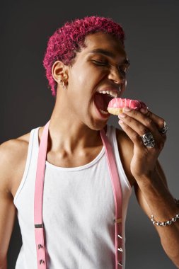 Pembe askılı genç adamın portresi. Pembe lezzetli donut yiyor. Moda ve tarz.