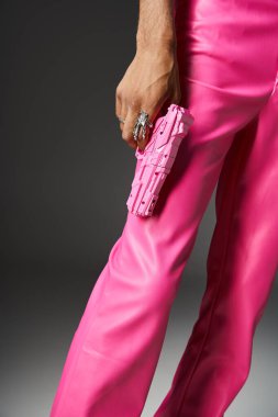 cropped view of man in pink leather pants with silver ring holding pink toy gun on gray backdrop clipart