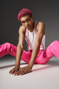 Pembe saçlı Afro-Amerikalı canlı giyinmiş yerde oturmuş kameraya bakıyor.
