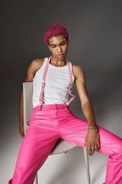 Pembe pantolonlu yakışıklı adam sandalyede gri arka planda poz veriyor, moda anlayışı.