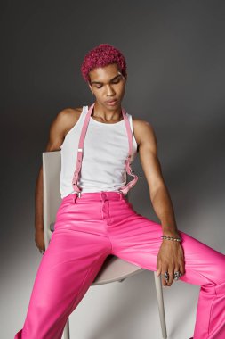 handsome stylish man in pink pants with suspenders posing on chair on gray backdrop, fashion concept clipart
