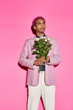 Pembe saçlı, elinde gül buketi olan yakışıklı erkek manken pembe arka plana bakıyor.