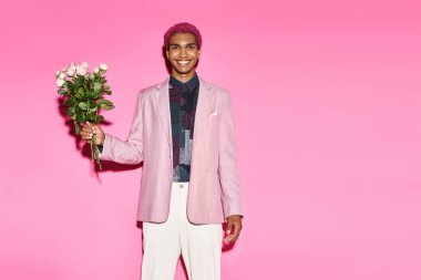 Pembe saçlı yakışıklı adam pembe arka planda gül buketi ile poz veriyor kameraya neşeyle gülümsüyor.