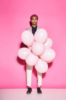 handsome man with pink hair holding pink balloons and looking at camera posing on pink backdrop clipart