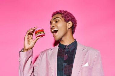 Kıvırcık pembe saçlı yakışıklı Afro-Amerikalı adam pembe arka planda mini burgerin tadını çıkarıyor.