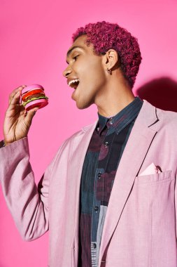 Pembe ceketli, gümüş küpeli, pembe arka planda poz veren, mini burger yiyen neşeli genç adam.