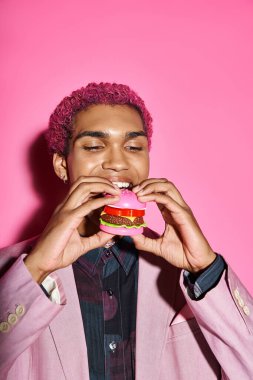 Güzel görünümlü, kıvırcık saçlı erkek model. Pembe arka planda lezzetli mini burgerler yiyor.
