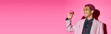 joyful handsome man with curly hair looking cheerfully at mini burger on pink background, banner clipart