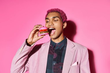 Gümüş küpeli şık Afrikalı Amerikalı adam pembe arka planda poz vermiş mini burger yiyor.