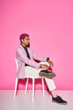 Pembe ceketli, kıvırcık saçlı şık adam pembe arka planda poz veren beyaz sandalyede doğal olmayan bir şekilde poz veriyor.