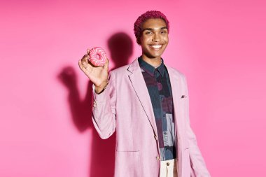 Canlı kıyafetli neşeli bir adam kameraya doğal olmayan bir şekilde gülümsüyor ve pembe arka planda donut tutuyor.