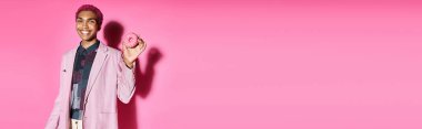 Canlı giyinmiş neşeli bir adam. Kameraya doğal olmayan bir şekilde gülümsüyor. Pembe arka planda donut tutuyor.