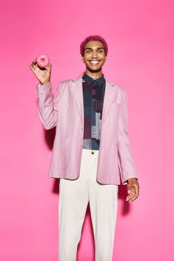 Yakışıklı Afro-Amerikan erkek model. Doğal olmayan bir şekilde gülümsüyor ve elinde donut tutuyor. Pembe arka plan.
