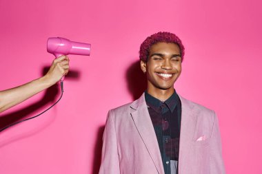 Kıvırcık saçlı, pembe saçlı, pembe arka fonda gözleri kapalı, elinde saç kurutma makinesi olan yakışıklı bir adam.