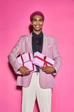 jolly african american man smiling weirdly at camera and holding presents in hands on pink backdrop clipart