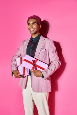 cheerful african american man in vibrant attire posing with presents in hands on pink backdrop clipart