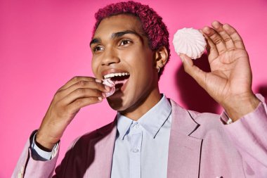handsome pink haired man in pink blazer eating zefir posing unnaturally posing on pink backdrop clipart