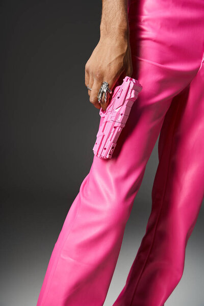 обрезанный вид человека в розовых кожаных штанах с серебряным кольцом держа розовый игрушечный пистолет на сером фоне