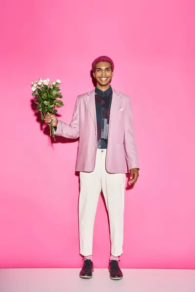 cheerful young man with bouquet of white roses posing on pink backdrop smiling sincerely at camera