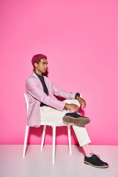 stylish man with curly hair in pink blazer posing unnaturally on white chair posing on pink backdrop