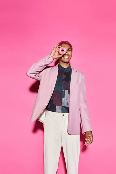 playful young man with curly pink hair posing with donut near his face posing on pink backdrop
