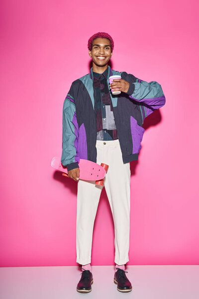 cheerful man in vibrant bright attire smiling and holding coffee and skateboard on pink backdrop